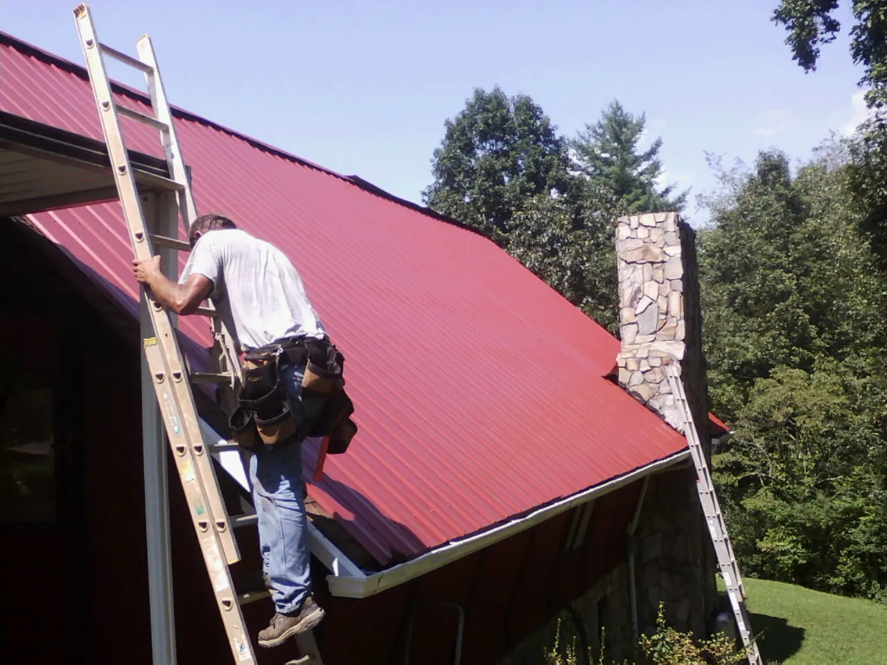 Roofer on a ladder beside a red metal roof and stone chimney — Soffit & Fascia Repair by Roofing Brothers in Juntura